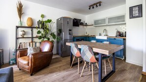Eine moderne Küche mit einer zentralen Kochinsel, ein Essbereich mit einem Holztisch und gemusterten Stühlen, ein Ledersofa und ein Bücherregal mit Dekorationselementen.