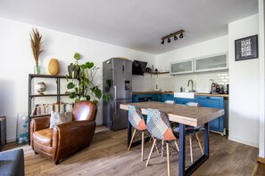 Una cocina moderna con una isla central, un área de comedor con una mesa de madera y sillas estampadas, un sofá de cuero y una estantería con objetos decorativos.
