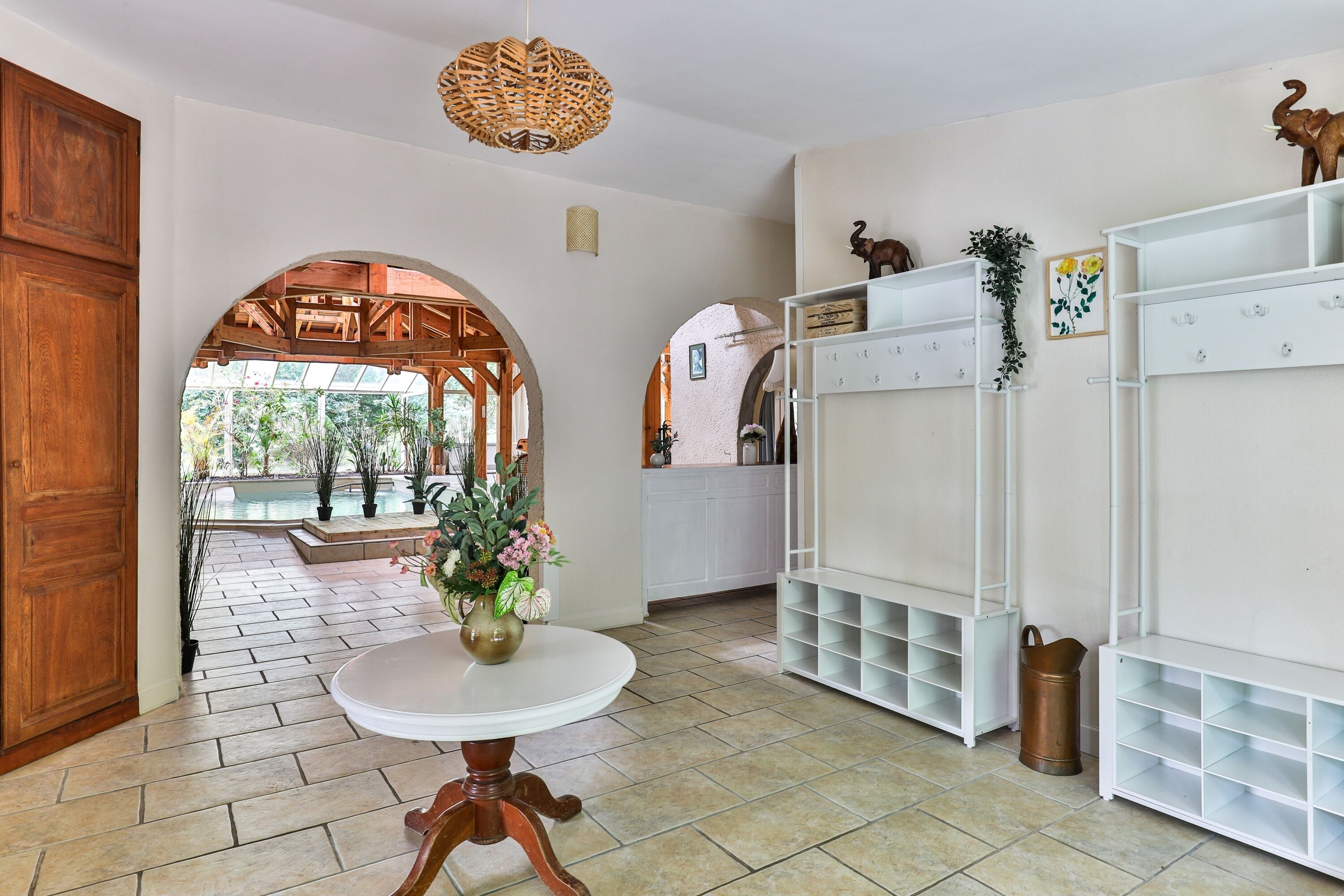 A spacious room with a large wooden door, a round table with a vase, white shelving units, and an arched doorway leading to a pool area.