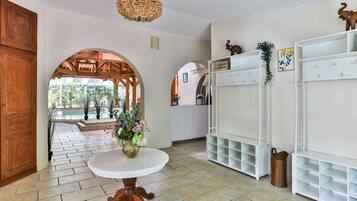 A spacious room with a large wooden door, a round table with a vase, white shelving units, and an arched doorway leading to a pool area.