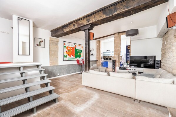 Uma sala de estar moderna com um sofá branco, uma televisão e uma cozinha ao fundo.