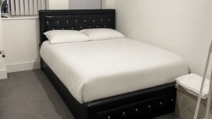 Une chambre à coucher bien rangée, avec un lit, une fenêtre équipée de stores, une table de chevet et une commode blanche.