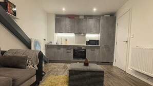 Un salon moderne avec un canapé, un pouf et une télécommande, donnant sur une cuisine aux meubles gris et équipée d’un four à micro-ondes.
