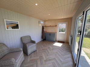 A room with a herringbone floor, a wicker chair, a small kitchen area, and a sliding glass door leading to an outdoor area. - Charming 3 Bedroom Cottage - Garden, WiFi, Parking (Holyhead)