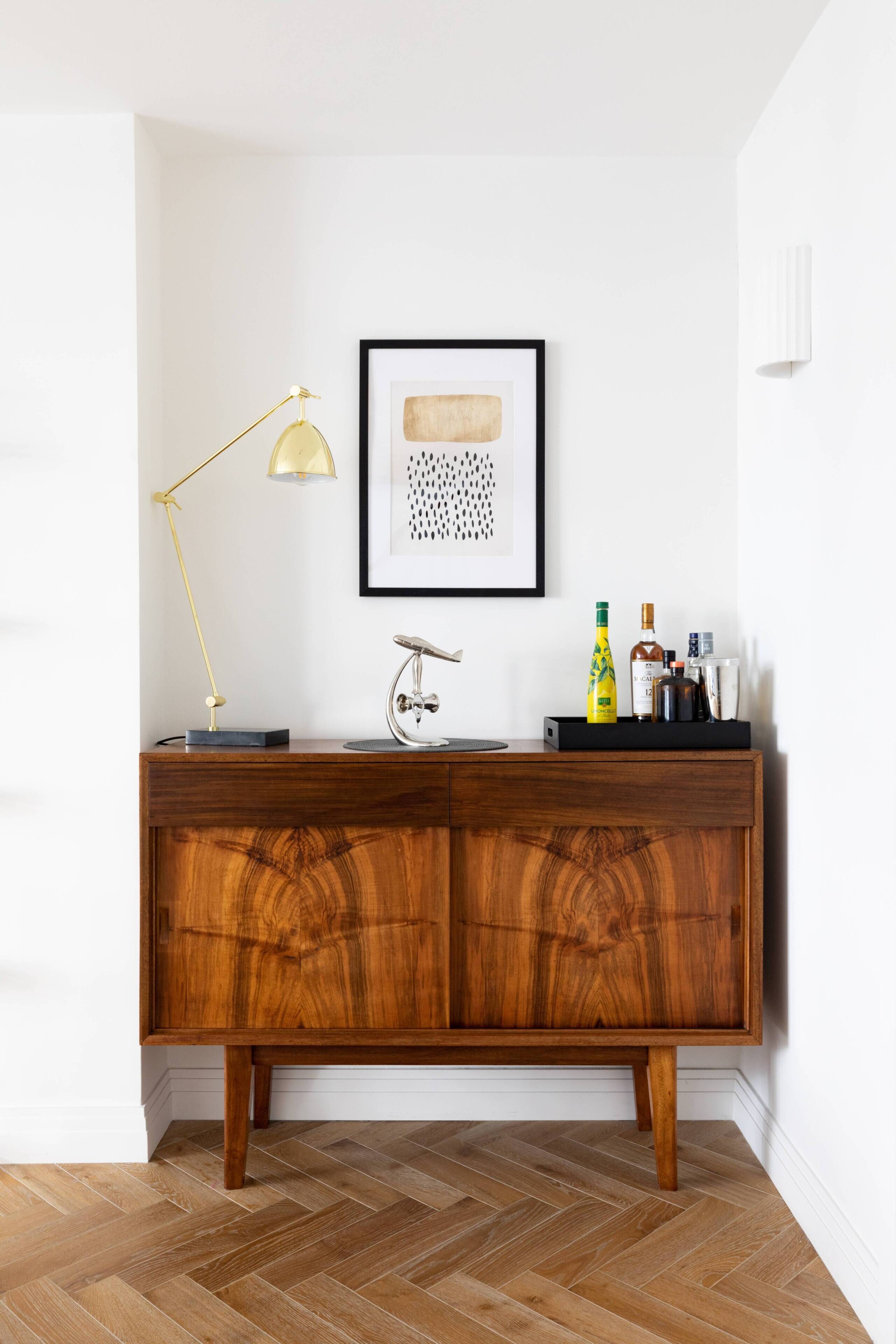 Un meuble en bois surmonté d’une lampe dorée, d’une œuvre d’art abstraite encadrée et d’une bouteille.