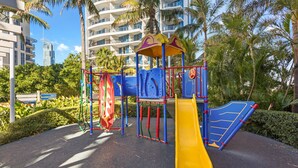Ein Spielplatz mit einer gelben Rutsche und Klettergerüsten, umgeben von Palmen und modernen Gebäuden.