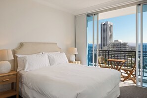 Habitación de hotel con una cama grande, una mesita de noche, una lámpara y un balcón con vistas a edificios altos.