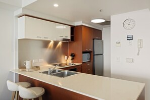 A modern kitchen with a stainless steel refrigerator, a microwave, and a sink. There are two bar stools and a knife block on the counter. - Chevron Renaissance River View (Surfers Paradise)