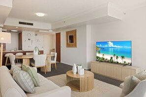 A modern living room with a large TV displaying a beach scene, a dining area with wooden furniture, and a cozy seating area with a round coffee table. - Chevron Renaissance River View (Surfers Paradise)