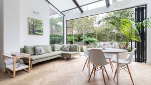 A modern living room with a sectional sofa, a dining table with chairs, a side table, and a large window with a view of greenery. - South End Green II - Five Bedroom House, Sleeps 10 (Hampstead)