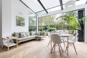 A modern living room with a sectional sofa, a dining table with chairs, a side table, and a large window with a view of greenery. - South End Green II - Five Bedroom House, Sleeps 10 (Hampstead)