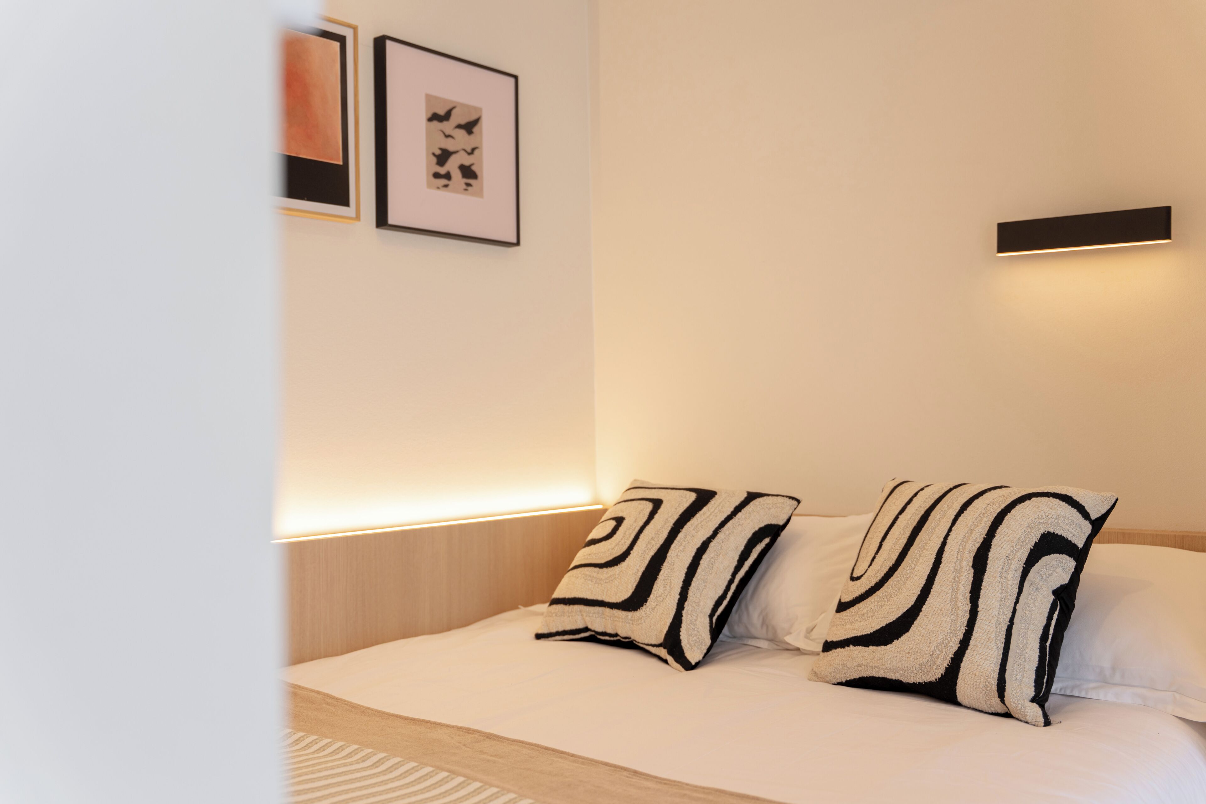 A modern hotel room with a neatly made bed, two pillows, a bedside table, and wall-mounted lighting.