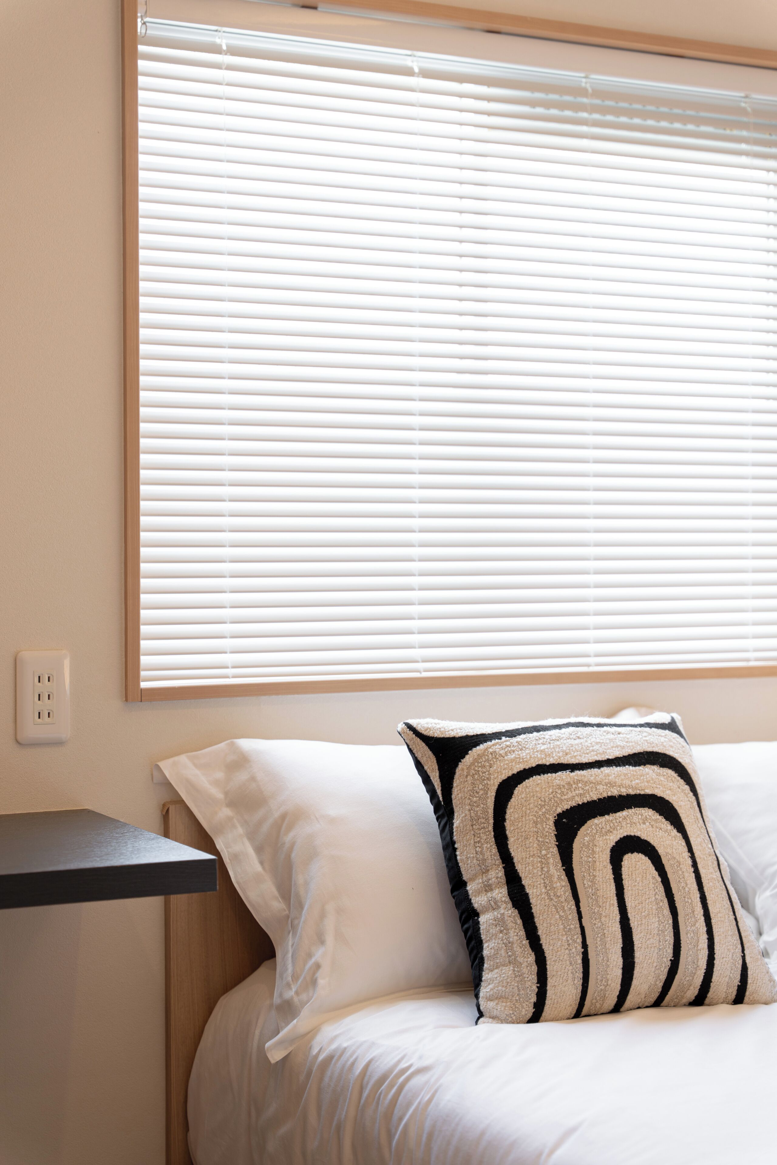 Un dormitorio con cama, ropa de cama blanca, almohada negra y beige, cabecero de madera y una ventana con persianas.