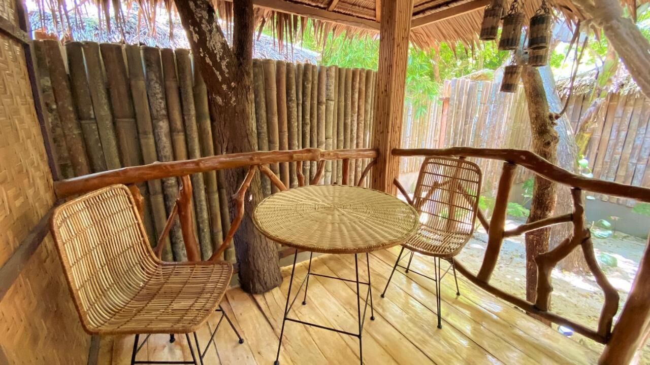 Una terraza de madera con una mesa redonda y dos sillas de mimbre, rodeada de paredes de bambú y un techo de paja.