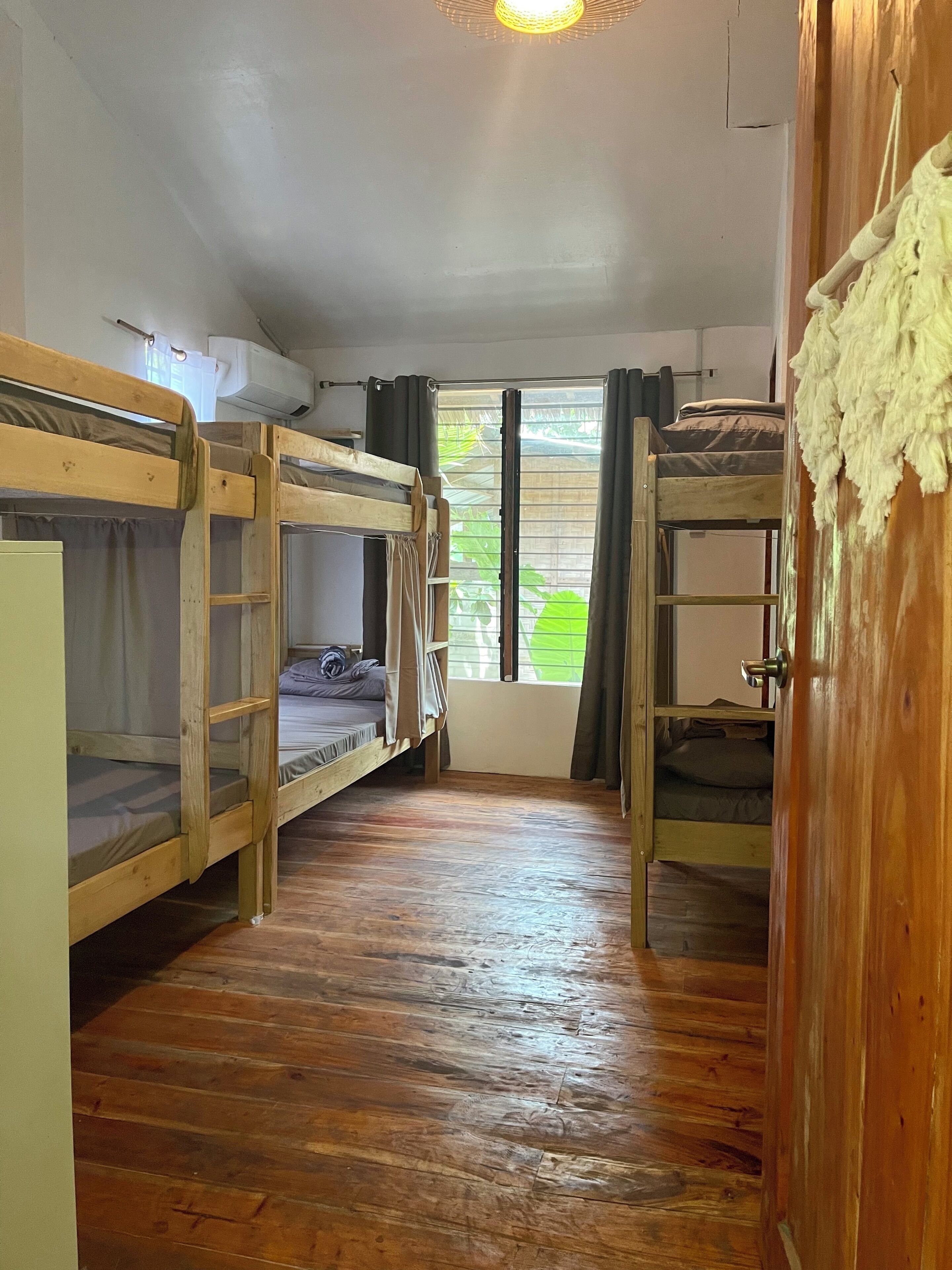 A room with bunk beds, a wooden floor, a window with curtains, and a door.