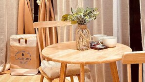 Una mesa de comedor de madera con sillas, un jarrón con flores, un juego de café y una bolsa de lavandería con la etiqueta 'WASH DRY IRON LAUNDRY DELUXE'.