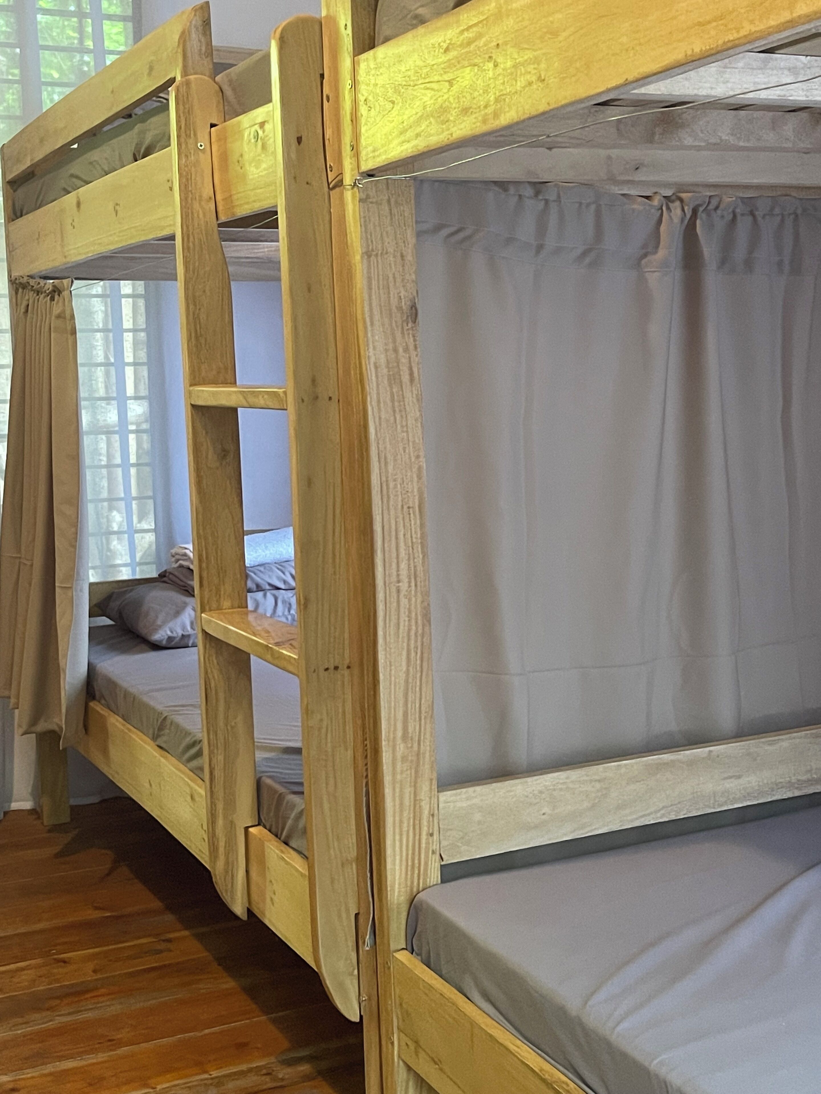 Una cama litera con estructura de madera, ropa de cama gris y una ventana con cortinas.