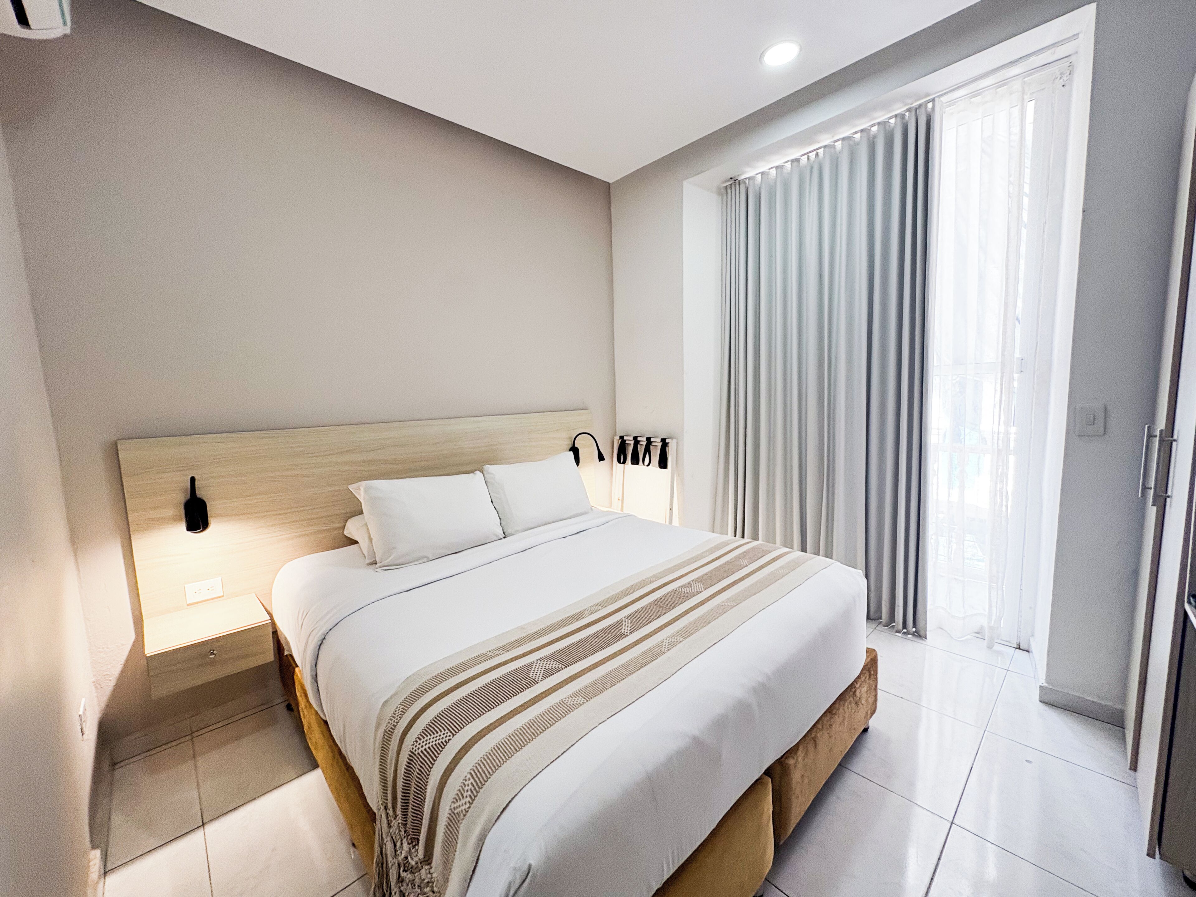 A hotel room with a bed, a nightstand, a wall-mounted lamp, a window with curtains, and a door.