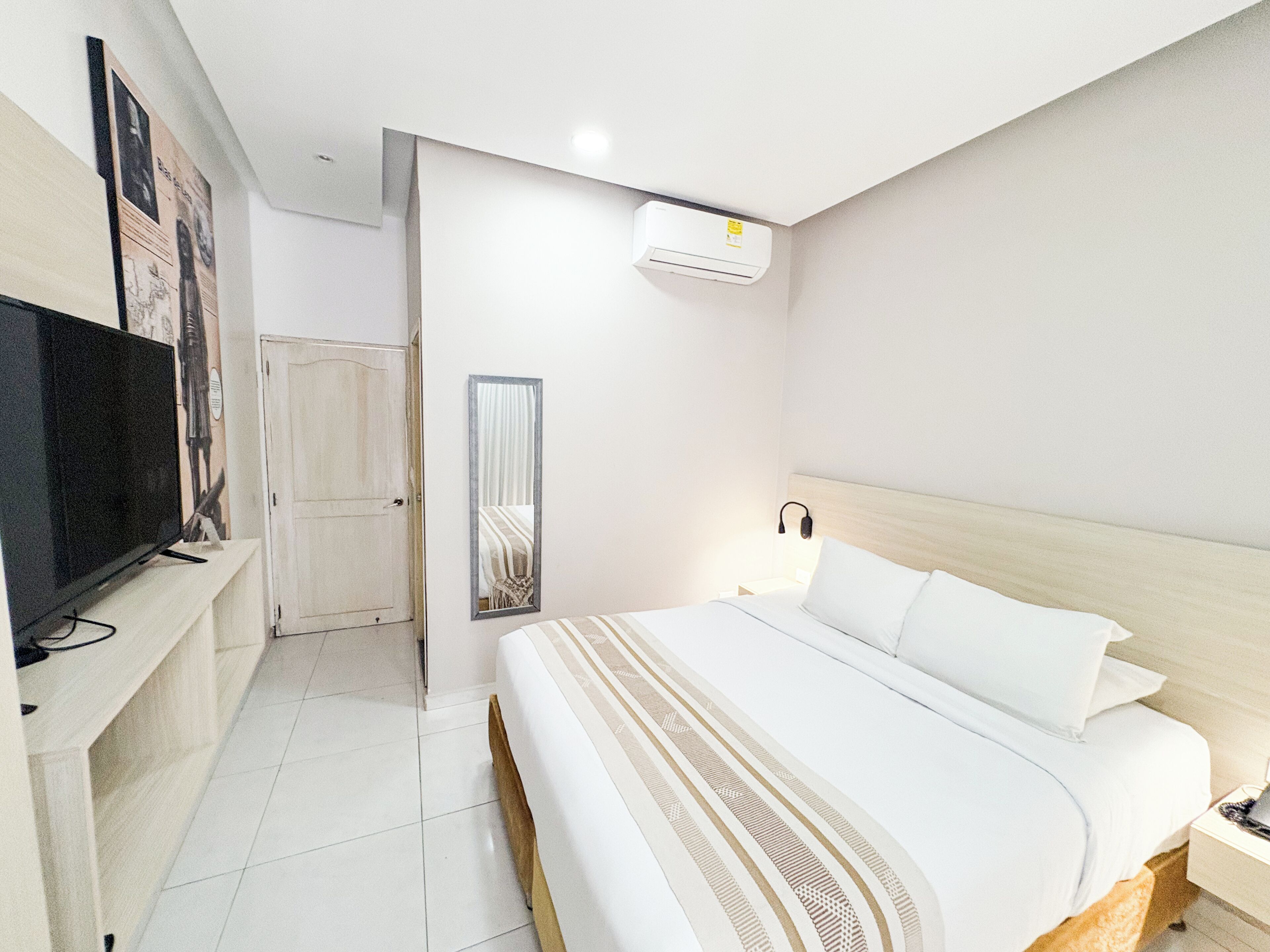 A hotel room with a bed, a TV, a mirror, and an air conditioning unit.