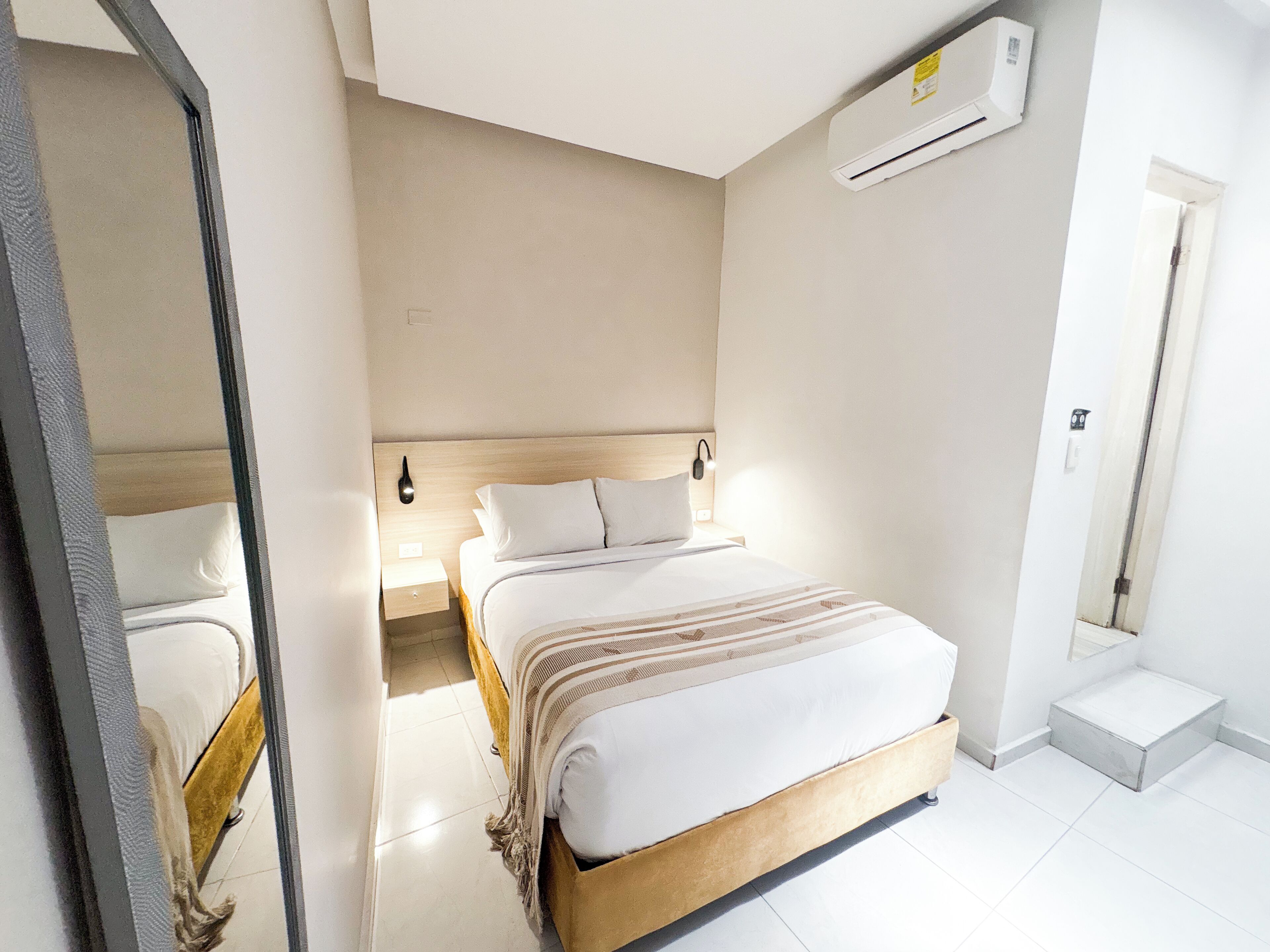 A hotel room with a bed, bedside tables, an air conditioning unit, and a door.