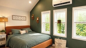 A bedroom with a bed, bedside tables, a nightstand, a wall-mounted air conditioner, and windows with hanging plants. -     1780 Post & Beam Studio - Historic Charm Near Downtown Keene (Keene)