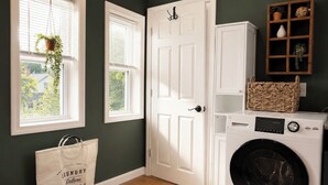 A laundry room with a washing machine, a door, a basket, and a bag. -     1780 Post & Beam Studio - Historic Charm Near Downtown Keene (Keene)