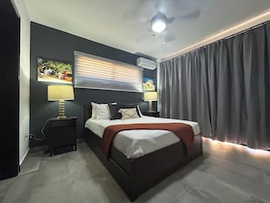 Ein Schlafzimmer mit einem Bett, zwei Nachttischen mit Lampen, einem Deckenventilator, einer Klimaanlage und einem großen Fenster mit Vorhängen.
