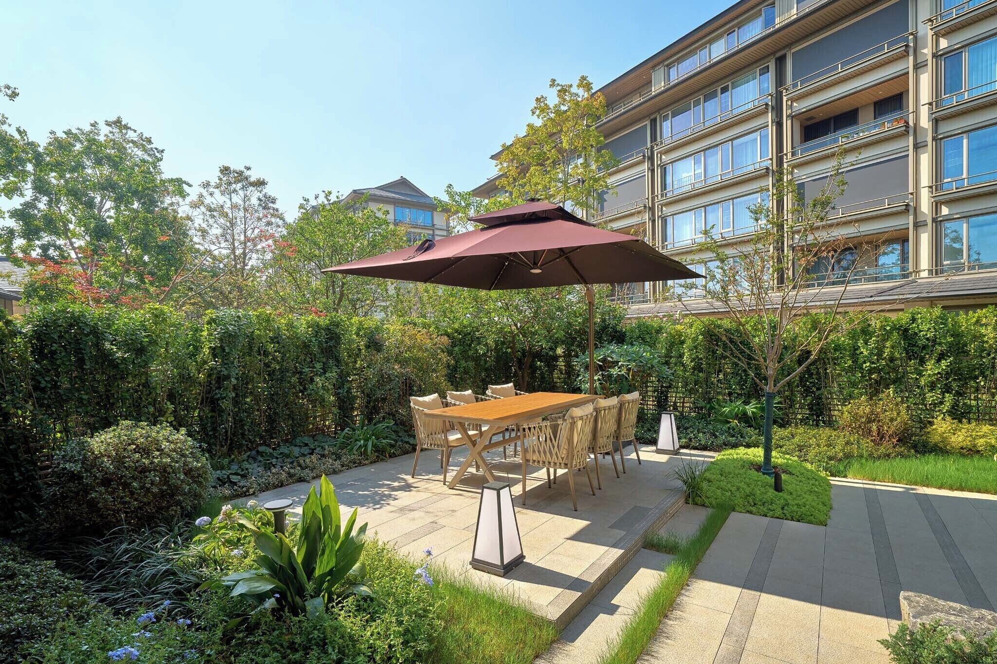 An outdoor patio with a wooden table and chairs, surrounded by lush greenery and a large umbrella.