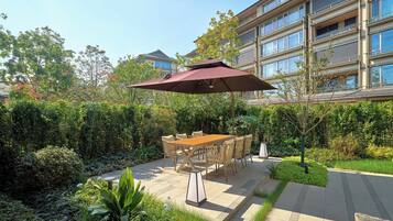 An outdoor patio with a wooden table and chairs, surrounded by lush greenery and a large umbrella.