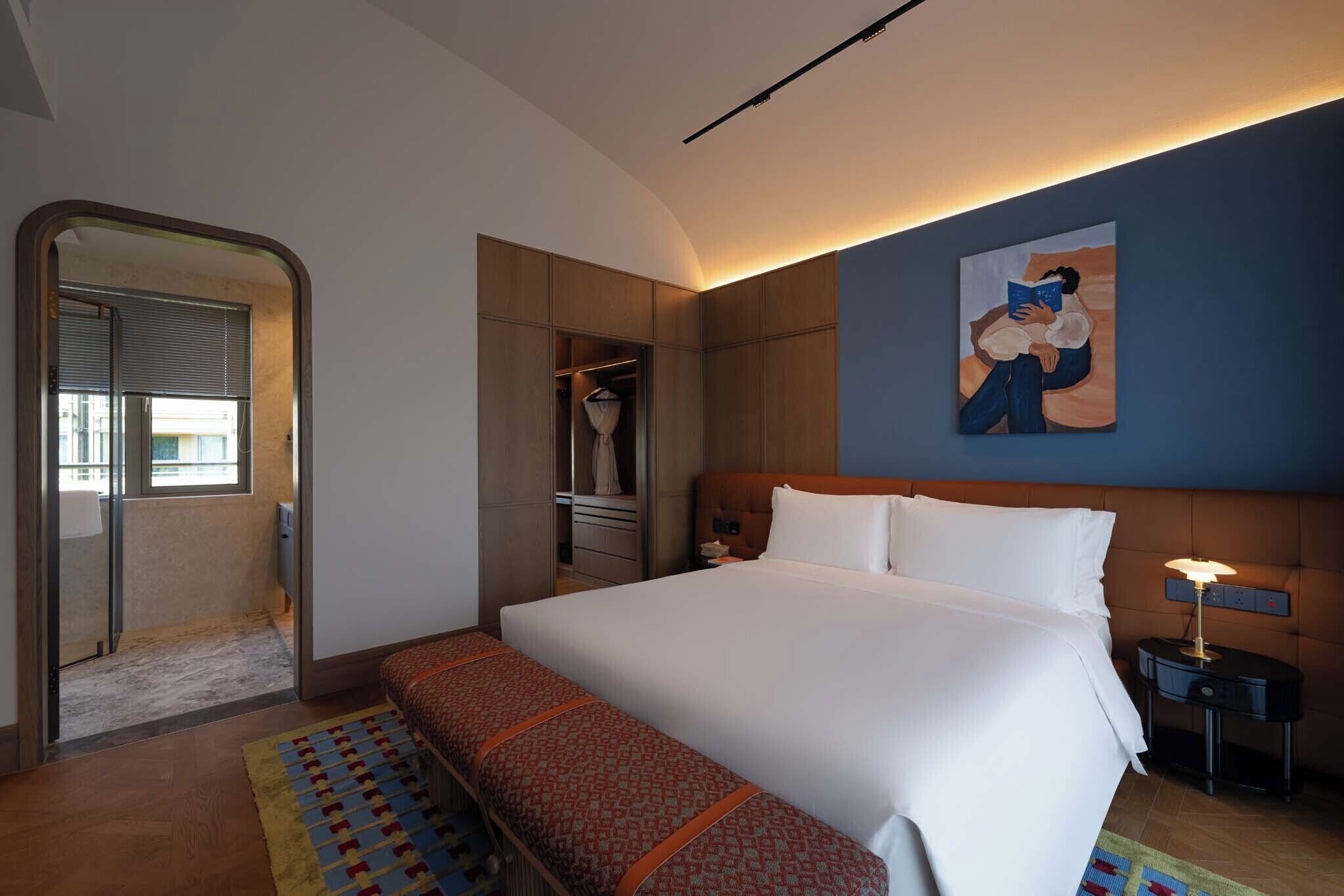 Habitación de hotel con cama, mesita de noche, lámpara, obra de arte, armario y vista a través de una puerta.