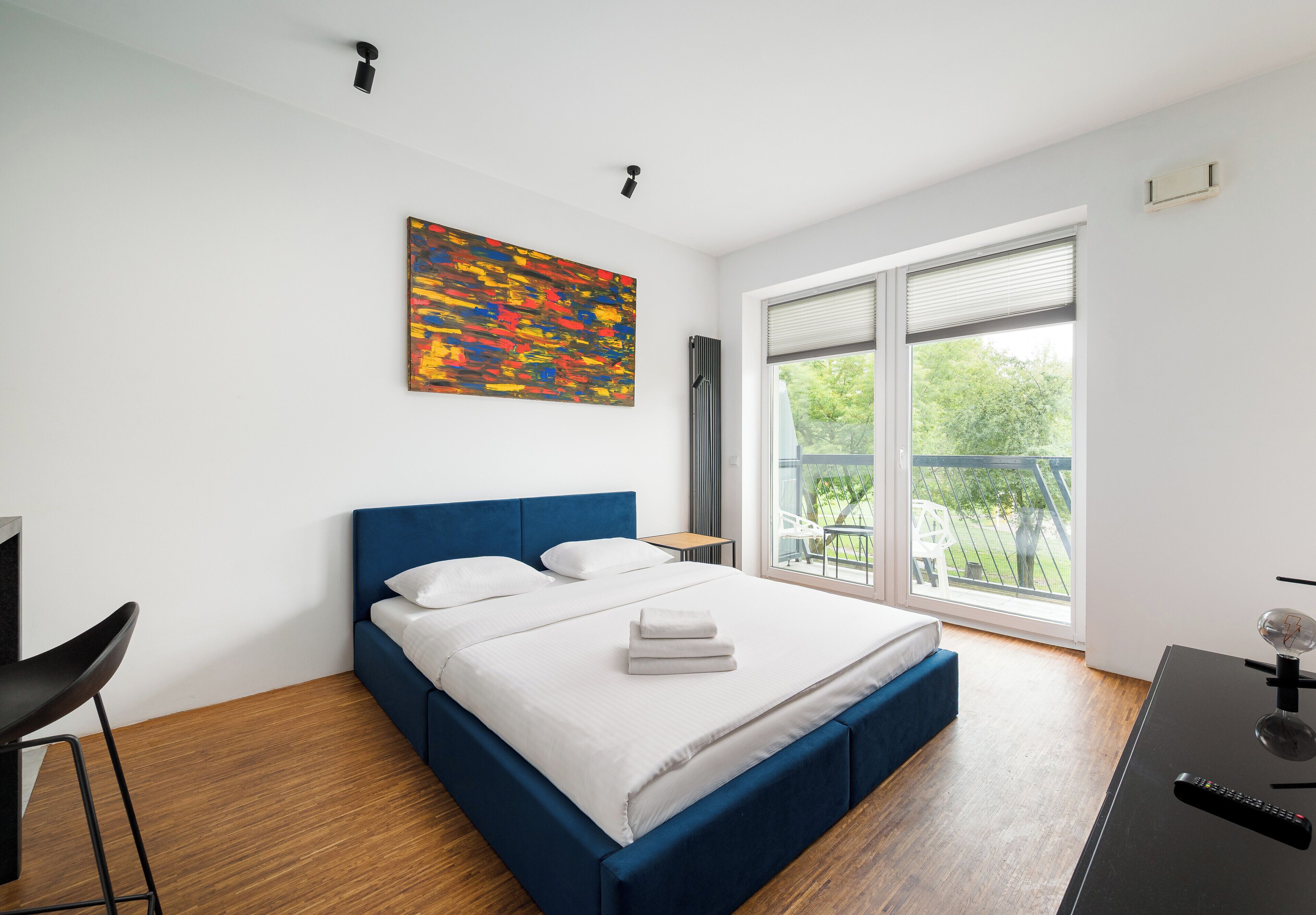 A modern bedroom with a large bed, a desk, and a window with a view of greenery.