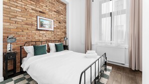 Een slaapkamer met een bakstenen muur, een bed met wit beddengoed, een nachtkastje met een lamp en een raam met gordijnen.