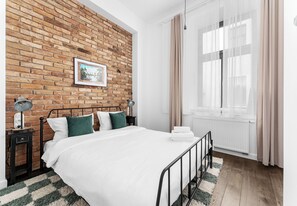 Ein Schlafzimmer mit Backsteinwand, einem Bett mit weißer Bettwäsche, einem Nachttisch mit Lampe und einem Fenster mit Vorhängen.