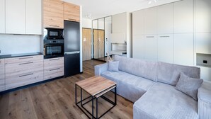Eine moderne Küche mit Holzschränken, einem schwarzen Kühlschrank und einer Mikrowelle. Im Wohnbereich befindet sich ein Holztisch und ein gestreiftes Sofa.