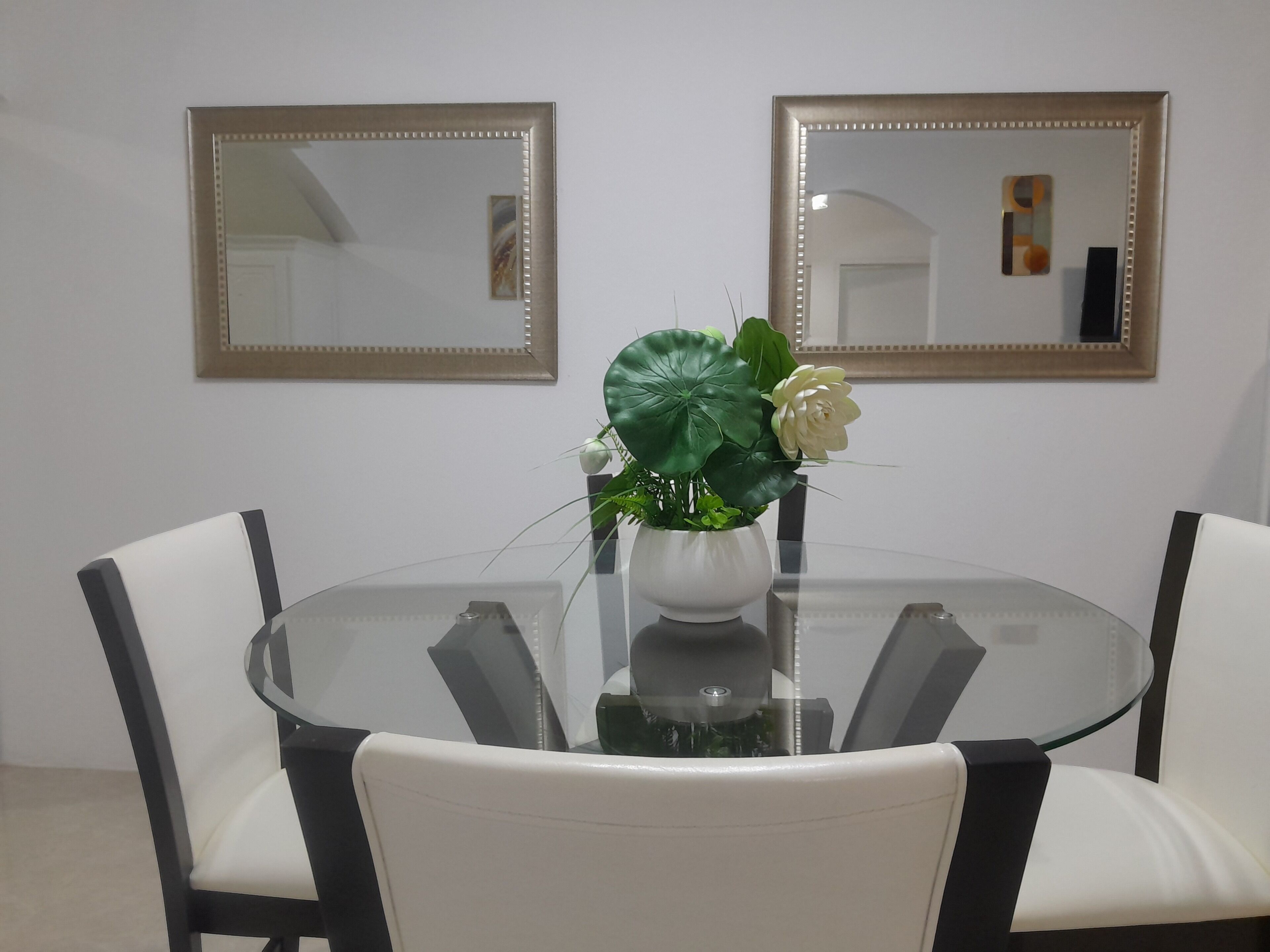Une table ronde en verre, sur laquelle repose un vase blanc contenant une plante et une fleur blanche, le tout entouré de chaises blanches.