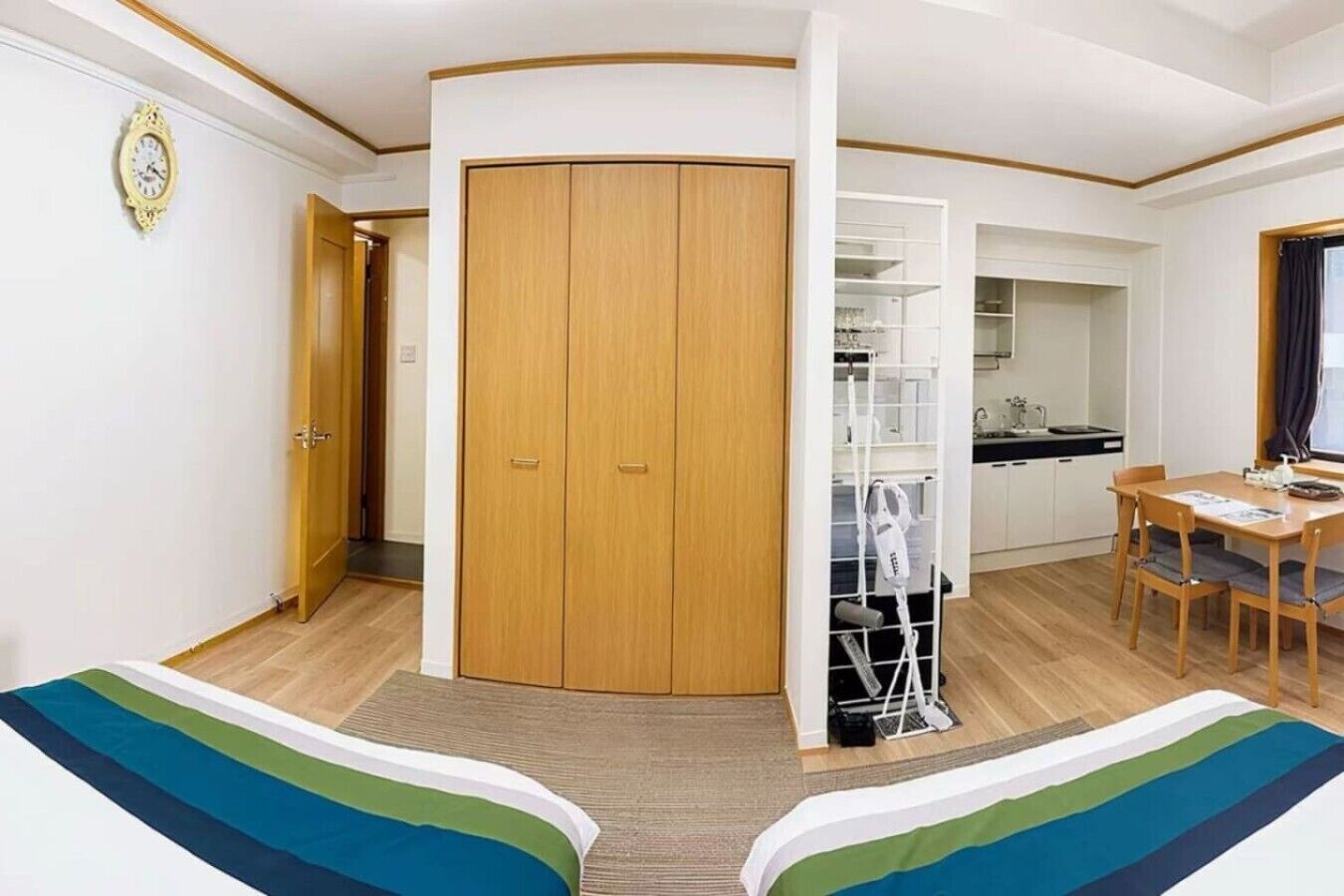 A room with a wooden wardrobe, a dining table with chairs, a kitchen area, and a bed with a striped bedspread.