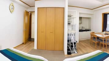 A room with a wooden wardrobe, a dining table with chairs, a kitchen area, and a bed with a striped bedspread.