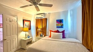 Quarto com cama, ventoinha no teto, mesa de cabeceira com abajur, porta-retrato e janela com cortinas.