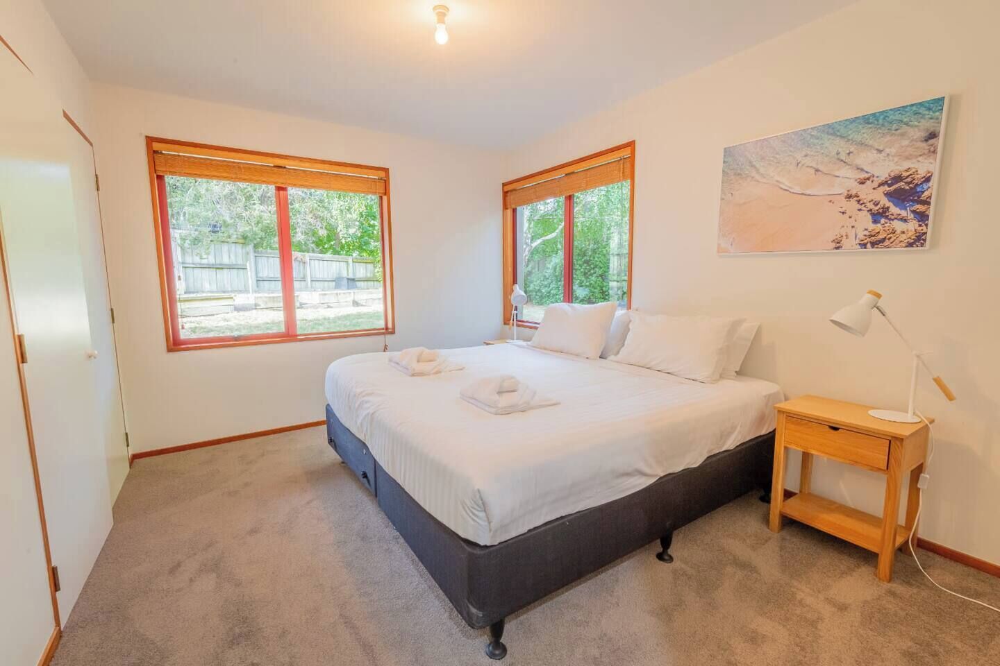 Une chambre à coucher avec un lit, deux fenêtres, une table de chevet, une lampe et un tableau au mur.