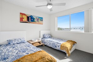 Una habitación con dos camas, un ventilador de techo, un cuadro de cabañas de playa y un ventanal con vistas.