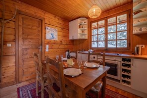 Interior de cabana de madeira com mesa de jantar posta para dois, área de cozinha com fogão e janela com vista para a neve.
