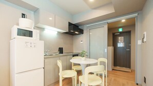 A compact kitchen with a refrigerator, microwave, and sink, a small dining table with chairs, and a door leading to an elevator. - Tora Hotel Nishikamata (Ota City)