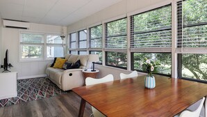 Una sala de estar moderna con una mesa de comedor de madera, sillas blancas, un sofá con cojines y ventanas amplias con persianas.