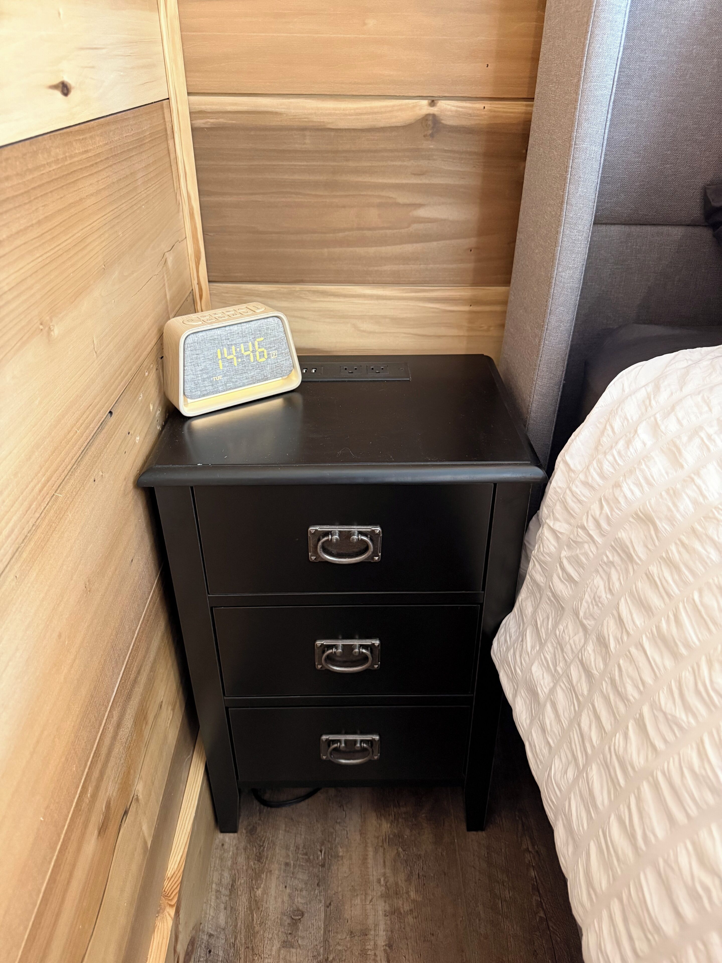 A wooden wall, a black nightstand with drawers, a digital clock, and a bed with a white quilt.
