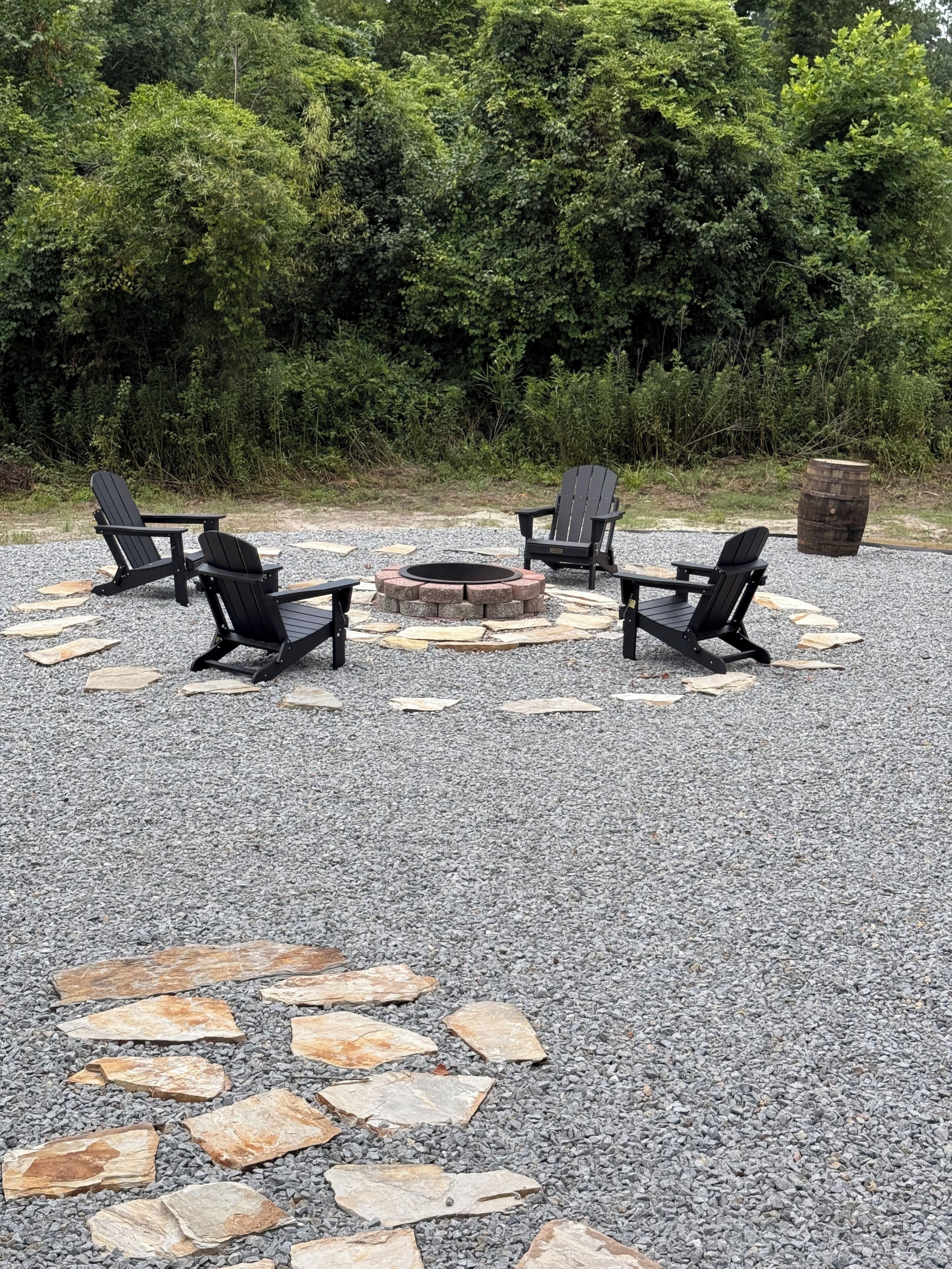Un patio de grava con cuatro sillas Adirondack, un pozo de fuego de piedra y un barril de madera.