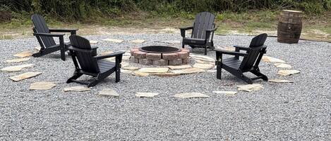 Un patio de grava con cuatro sillas Adirondack, un pozo de fuego de piedra y un barril de madera.
