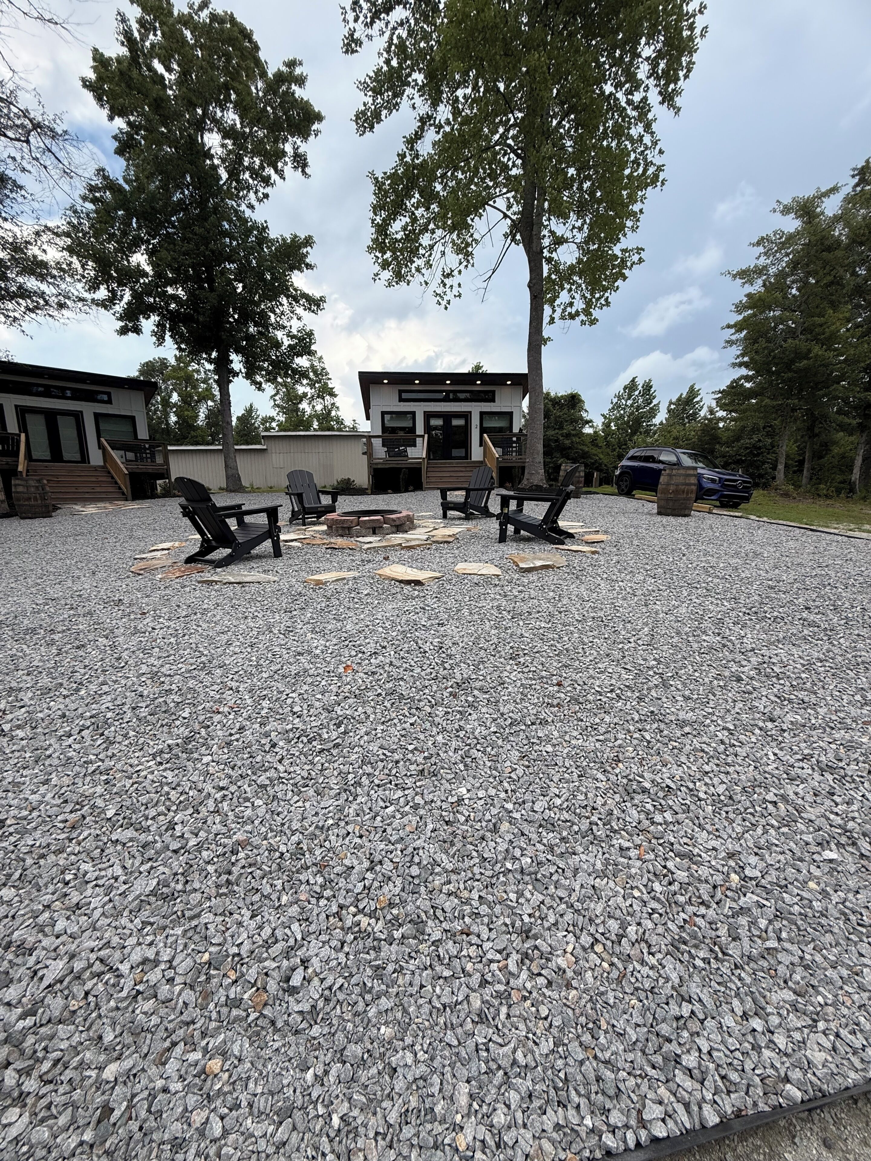 A modern outdoor seating area with a fire pit, surrounded by gravel, near a building with a deck and a car parked nearby.