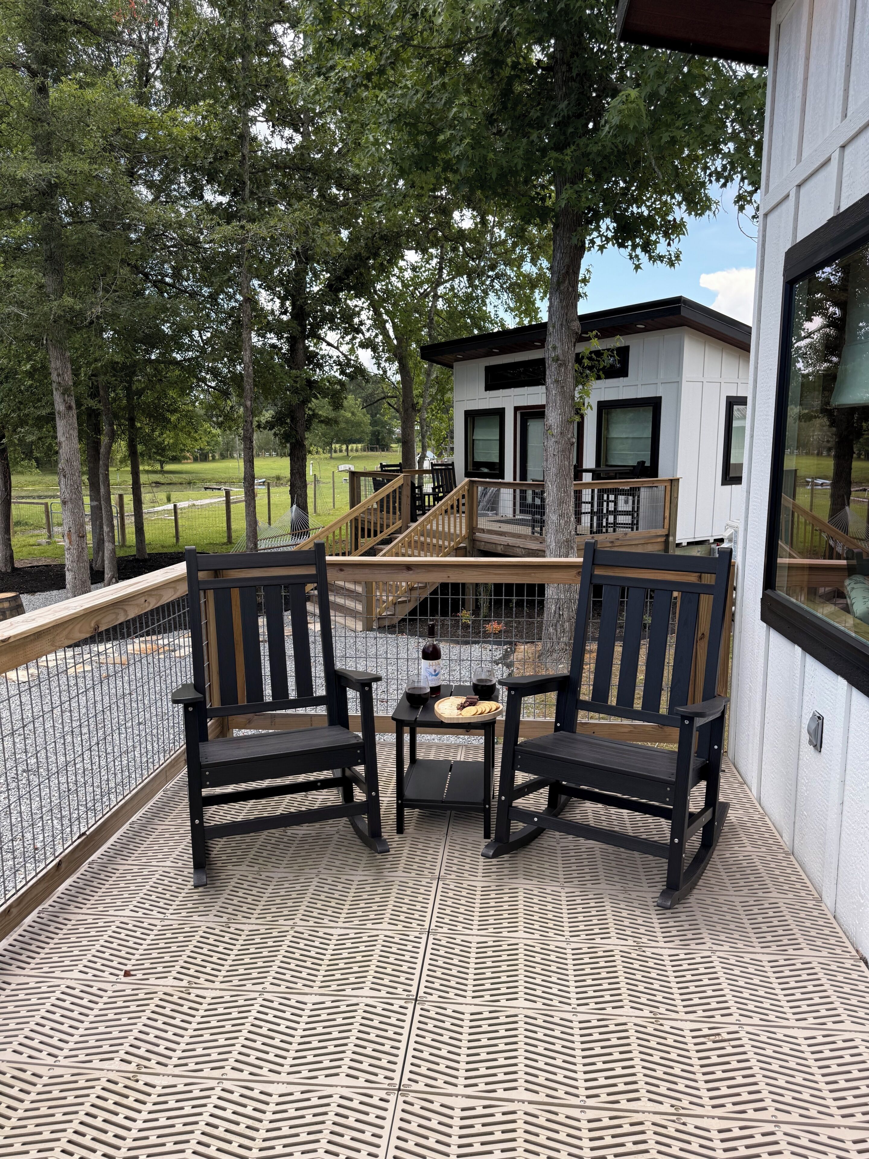 A covered outdoor seating area with two chairs and a table, a small deck with a railing, and a view of a building and trees in the background.