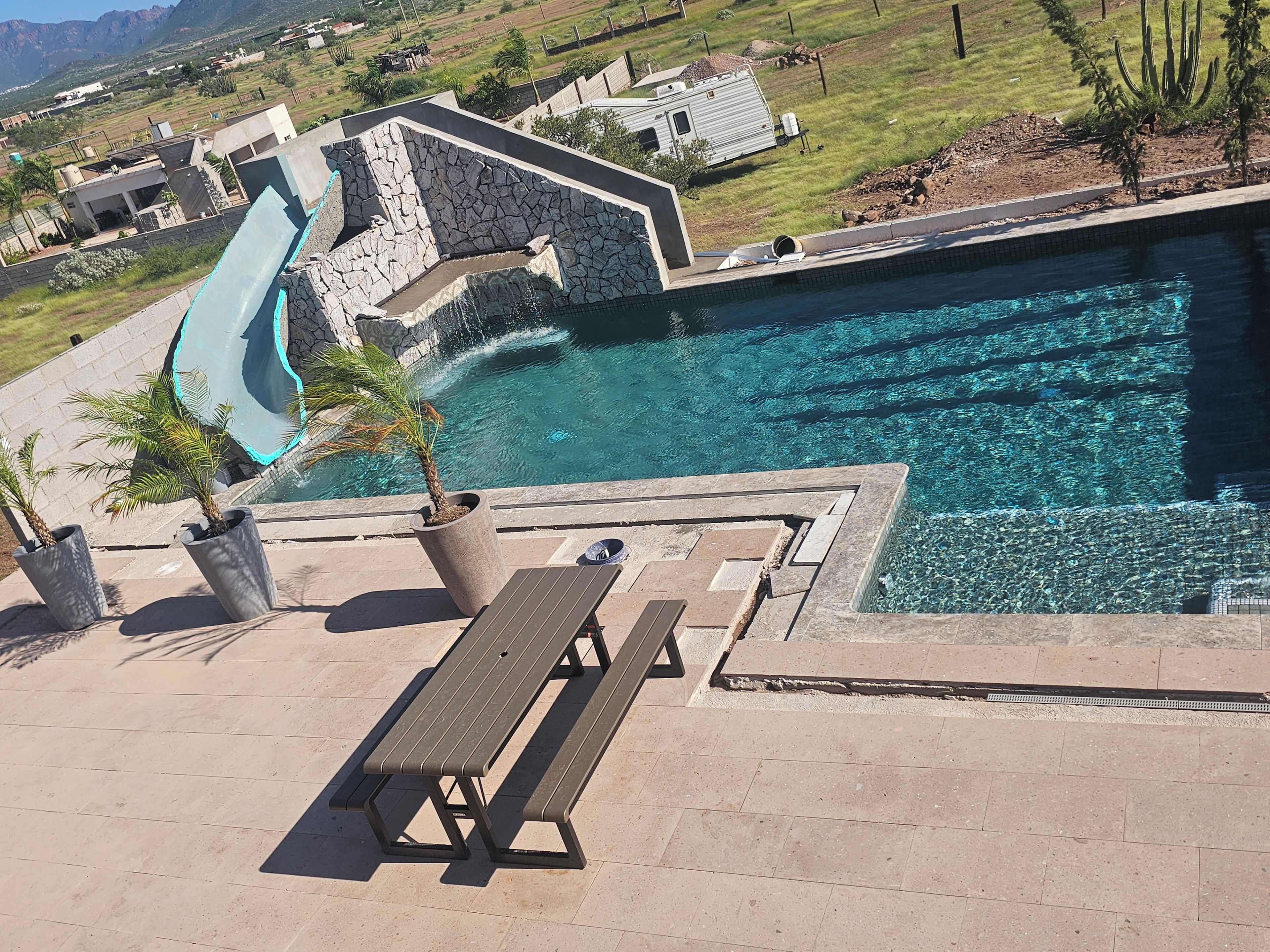 A swimming pool with a slide, surrounded by a stone wall and a picnic table.