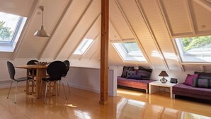 Um quarto com pé-direito alto e janelas de iluminação natural, área de jantar com cadeiras, sofá e uma pequena mesa.